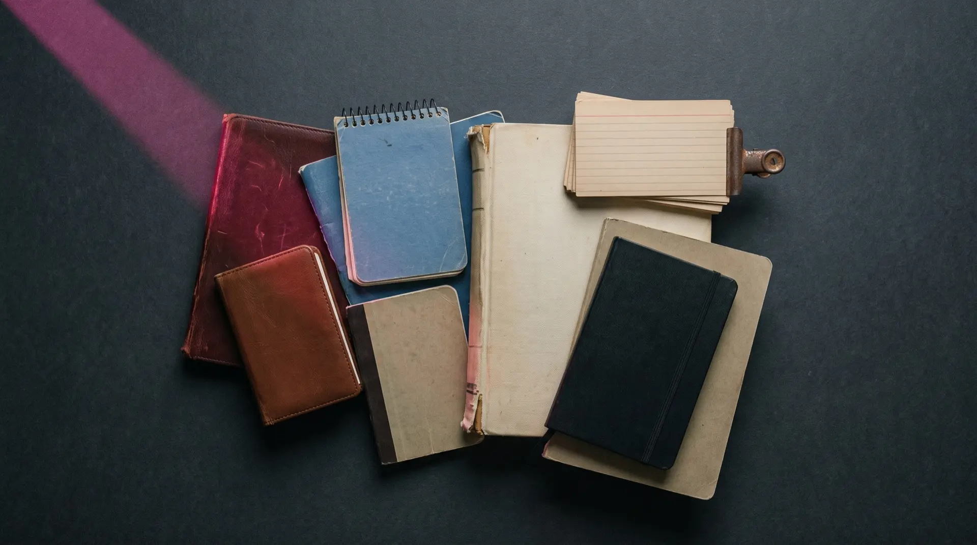 A pile of worn notebooks, pocket pads and a clip of ruled index cards on a dark surface